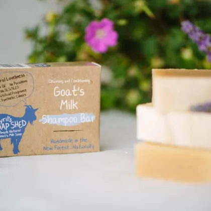 Cyrils soap shed shampoo bar stacked with other bars to the right with the brown cardboard box on the left a green and purple arrangment is in the background