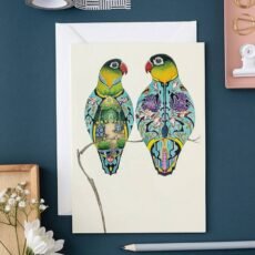 Lovebirds greetings card by The DM Collection with it's envelope on a dark blue table with a pencil some flowers and a bulldog clip