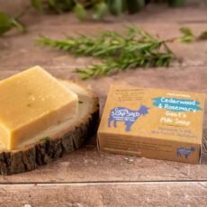 Cedarwood and rosemary goats milk soap bar displayed on a slice of wood with the box next to it with a sprig of rosemary in the background