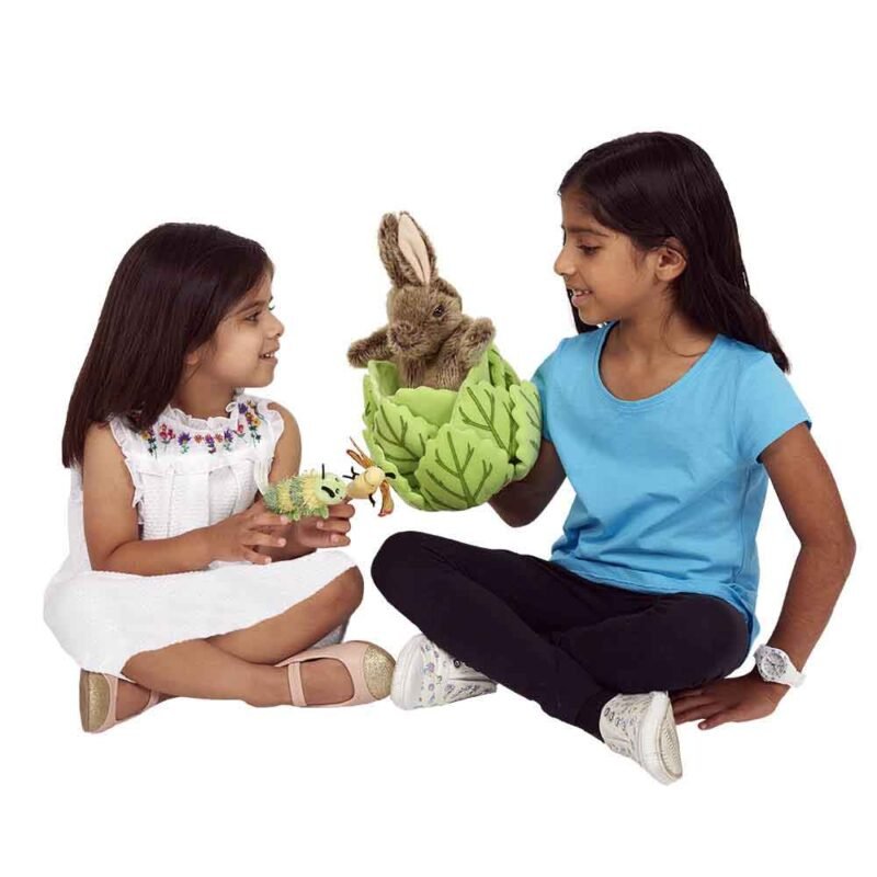2 young girls one in a white dress, the other in a blue top and trousers sit cross legged. The girl in blue holds the rabbit in lettuce hand puppet while the girl in white is looking on while wearing the caterpillar and butterfly finger puppets