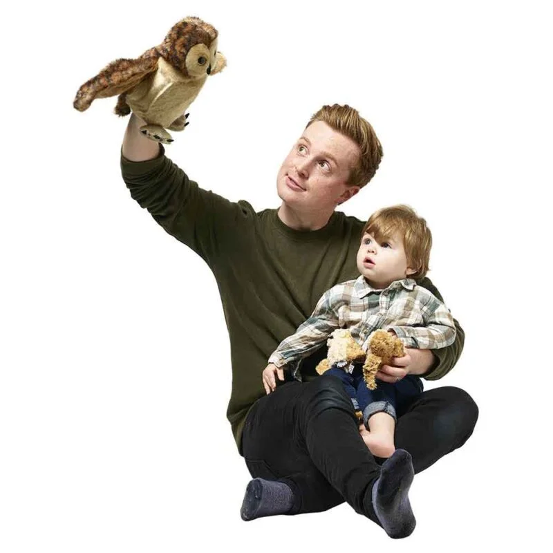 A man is sat ont he floor playing with the Tawny Owl hand puppet as though it is flying while a young child sitting on his lap is watching, the man holds an owlet finger puppet while another is on the childs lap