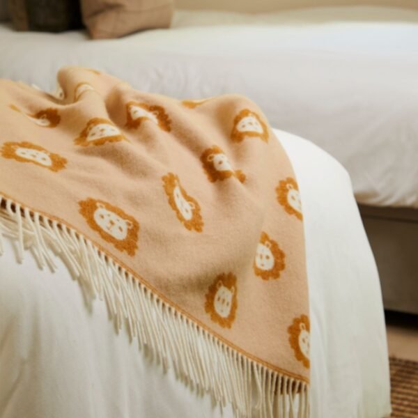 Merino wool baby blanket brown and cream with little cartoon lion faces and with a fringe laid over a light coloured bed spread