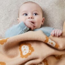 Baby in a grey jumper lying under a Merino wool baby blanket in brown and cream with cartoon lion faces