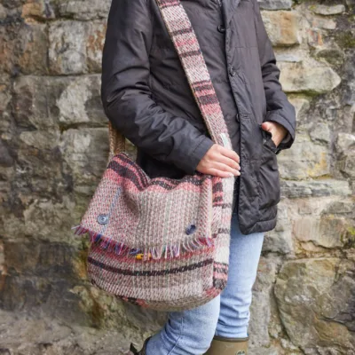 random recycled wool bag worn over the shoulder by a woman in jeans and dark coat against a stone wall